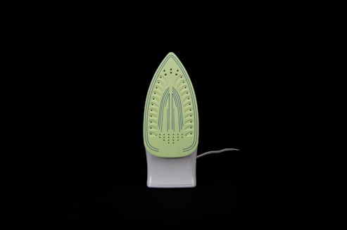 An iron is placed upright against a black background. The soleplate is visible and has a light green color with blue stripe details and multiple steam holes.
