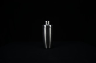 A stainless steel cocktail shaker is centered against a dark background. The shaker has a smooth, reflective surface and tapers towards the bottom.