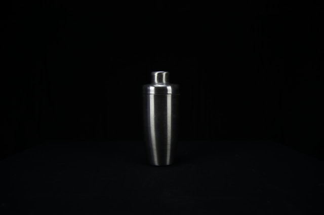 A stainless steel cocktail shaker is centered against a dark background. The shaker has a smooth, reflective surface and tapers towards the bottom.