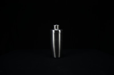 A stainless steel cocktail shaker is centered against a dark background. The shaker has a smooth, reflective surface and tapers towards the bottom.
