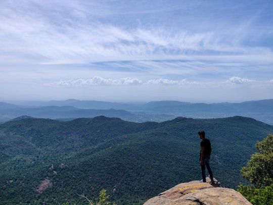 man wearing black t-shirt standing on rocky hill viewing mountainunder blue and white sky in Yercaud India