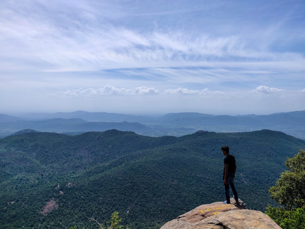 man wearing black t-shirt standing on rocky hill viewing mountainunder blue and white sky