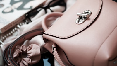 A close-up of a light pink leather handbag with a silver clasp, accompanied by a pair of glasses, a book, a scrunchie, and pearls. The items are arranged on a soft, light-colored fabric, contributing to an elegant and feminine aesthetic.