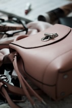 A pink leather handbag is prominently placed in the foreground with its metal clasp in focus. Surrounding the handbag are blurred items such as sunglasses with a black frame, makeup products including a lipstick, and some rolled-up magazines. The scene emits an elegant and stylish atmosphere.