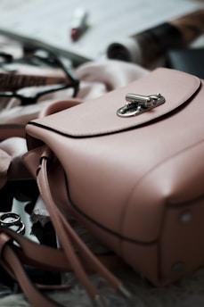 A pink leather handbag is prominently placed in the foreground with its metal clasp in focus. Surrounding the handbag are blurred items such as sunglasses with a black frame, makeup products including a lipstick, and some rolled-up magazines. The scene emits an elegant and stylish atmosphere.