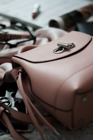 A pink leather handbag is prominently placed in the foreground with its metal clasp in focus. Surrounding the handbag are blurred items such as sunglasses with a black frame, makeup products including a lipstick, and some rolled-up magazines. The scene emits an elegant and stylish atmosphere.