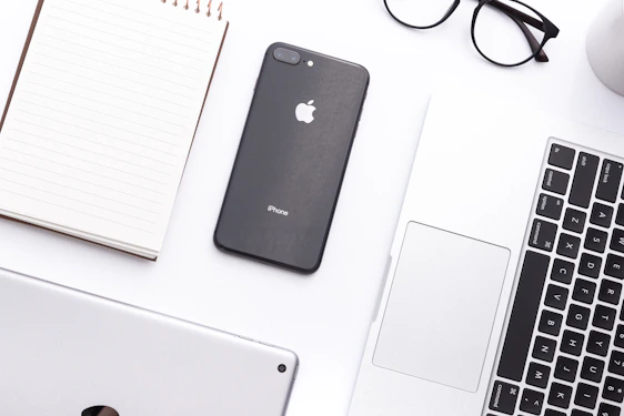A sleek, minimalist workspace featuring a MacBook, iPhone, and iPad arranged on a clean white desk with soft natural light.