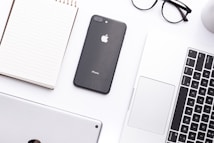 A neatly arranged workspace features a notepad, a black iPhone, a silver laptop with a visible keyboard, and a pair of black-framed glasses. The setup is on a clean white surface, creating a minimalist and organized atmosphere.