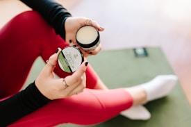 A person sits on a green yoga mat, wearing bright pink leggings and white socks. They hold two small jars, one with a green label reading 'CBD Balm' and the other lid removed, showing its contents. The person wears rings and has black-painted nails.