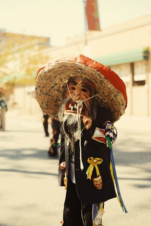 A person is wearing a colorful, traditional costume with a large, decorated straw hat and a detailed mask portraying a bearded face. The outfit includes various colorful patches and ribbons. They appear to be participating in a cultural or festive event outdoors, as sunlight casts shadows on the pavement.