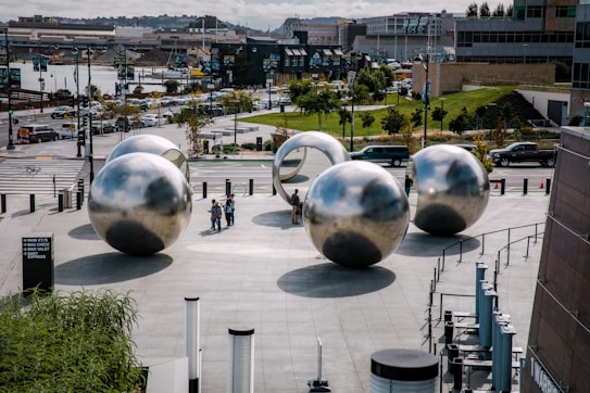 A public urban space features several large reflective metal spheres arranged artistically on a plaza. People walk among the spheres, interacting and observing the installation. In the background, a street with vehicles and a landscaped area with trees and grass can be seen, along with buildings and a body of water.