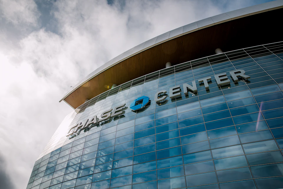 The image shows a large glass building with a curved roof, standing tall against a clear blue sky. The building prominently displays the Chase Center logo on its facade, indicating that it is likely an office or commercial space within the center. The perspective of the photo is from below, looking up at the building, which emphasizes its height and grandeur.

The sky above the building is filled with clouds, suggesting a bright and sunny day. The sun's rays are shining down on the building, highlighting its architectural features and making it stand out against the backdrop of the clear blue sky.