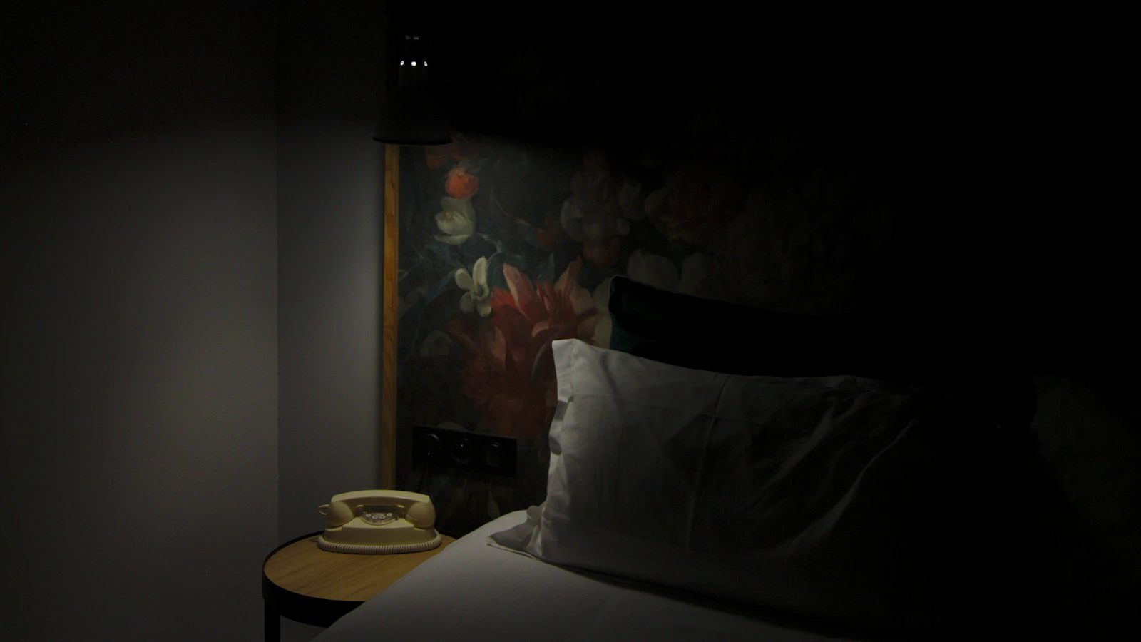 Photograph showing dark hotel room holds a made bed and a telephone on the bedside table.