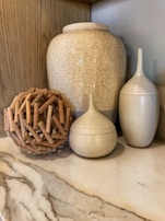 A decorative arrangement featuring a large ceramic vase with a crackled texture, accompanied by two smaller, similarly styled jars. Beside them is a spherical object made of intertwined wooden sticks. The setting is on a marble countertop with a beige wooden backdrop, creating a harmonious and earthy aesthetic.