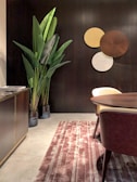 A modern interior space featuring a dark wood wall with decorative round panels in shades of brown, yellow, and white. A tall plant with large green leaves is placed next to a dark wooden cabinet on the left. A round wooden table and a chair with a cream-colored seat are visible, and the floor is covered by a striped textured rug in shades of pink and cream.