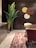 A modern interior space featuring a dark wood wall with decorative round panels in shades of brown, yellow, and white. A tall plant with large green leaves is placed next to a dark wooden cabinet on the left. A round wooden table and a chair with a cream-colored seat are visible, and the floor is covered by a striped textured rug in shades of pink and cream.