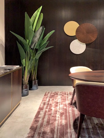 A modern interior space featuring a dark wood wall with decorative round panels in shades of brown, yellow, and white. A tall plant with large green leaves is placed next to a dark wooden cabinet on the left. A round wooden table and a chair with a cream-colored seat are visible, and the floor is covered by a striped textured rug in shades of pink and cream.