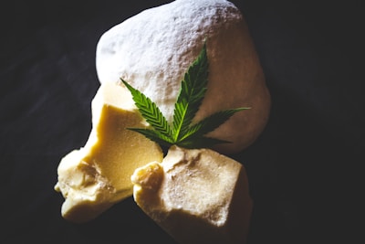 A green cannabis leaf is placed on top of two solid, uneven chunks of yellowish butter or wax-like substance and a larger white, fluffy powdery item. The background is dark, enhancing the composition's contrast.