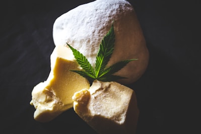 A green cannabis leaf is placed on top of two solid, uneven chunks of yellowish butter or wax-like substance and a larger white, fluffy powdery item. The background is dark, enhancing the composition's contrast.