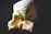 A green cannabis leaf is placed on top of two solid, uneven chunks of yellowish butter or wax-like substance and a larger white, fluffy powdery item. The background is dark, enhancing the composition's contrast.