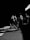 Silhouetted runners in a dramatic black and white scene, casting elongated shadows across a textured road surface. The high contrast highlights their dynamic movement and athletic form.