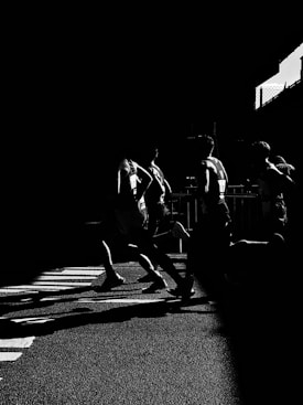 Silhouetted runners in a dramatic black and white scene, casting elongated shadows across a textured road surface. The high contrast highlights their dynamic movement and athletic form.
