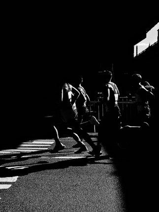 Silhouetted runners in a dramatic black and white scene, casting elongated shadows across a textured road surface. The high contrast highlights their dynamic movement and athletic form.