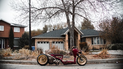 A modified motorcycle is parked on a suburban street in front of a house with a brick exterior and a double garage. The motorcycle has a unique design with a maroon frame and gold-rimmed wheels. Surrounding the area are bare trees and bushes, indicating a fall or winter setting.