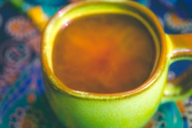 A close-up view of a green ceramic cup filled with a warm brown liquid, possibly tea or coffee. The cup is placed on a vibrant, patterned surface featuring a mix of swirling colors and intricate designs.