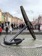 A large anchor sculpture is prominently displayed on a cobblestone surface in a busy public area. The surroundings include colorful, historic buildings in various shades, with people walking and engaging in various activities. Flags and lampposts are visible, adding to the lively, urban atmosphere.