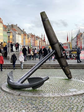 A large anchor sculpture is prominently displayed on a cobblestone surface in a busy public area. The surroundings include colorful, historic buildings in various shades, with people walking and engaging in various activities. Flags and lampposts are visible, adding to the lively, urban atmosphere.