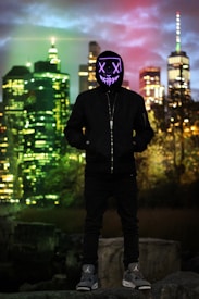 A person wearing a black hoodie and sneakers stands in front of an urban night skyline. Their face is obscured by a glowing LED mask with crossed eyes and a stitched mouth design. The background features illuminated skyscrapers under a cloudy sky displaying hues of green and red.