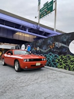 An orange sports car is parked on a gray brick surface in front of a colorful graffiti wall. The graffiti features abstract shapes and designs, including butterflies and faces. Above, an overpass is painted a light purple, with green highway signs indicating directions to Brooklyn Bridge and Manhattan Civic Center.