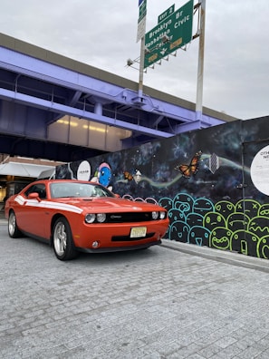 An orange sports car is parked on a gray brick surface in front of a colorful graffiti wall. The graffiti features abstract shapes and designs, including butterflies and faces. Above, an overpass is painted a light purple, with green highway signs indicating directions to Brooklyn Bridge and Manhattan Civic Center.