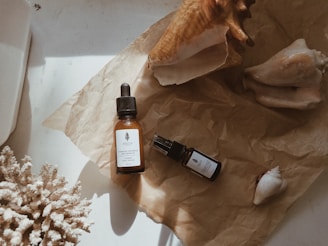 On a crumpled sheet of brown paper, there are two bottles of skincare products, one with a dropper, and various large seashells. A piece of coral is positioned on the left side.