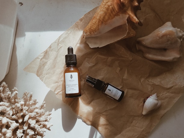 On a crumpled sheet of brown paper, there are two bottles of skincare products, one with a dropper, and various large seashells. A piece of coral is positioned on the left side.