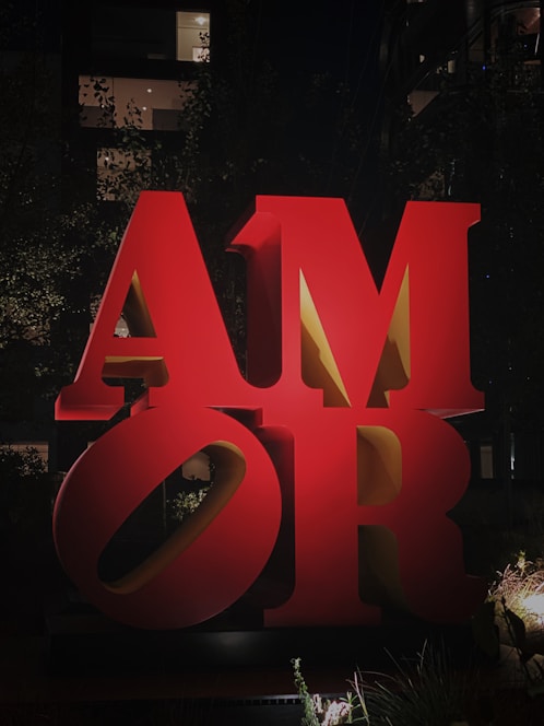 Large, three-dimensional red letters spelling out 'AMOR' are prominently displayed outdoors. The backdrop is dark, with a few illuminated windows and silhouetted trees visible, adding to the nighttime atmosphere.