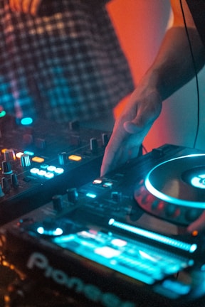 A DJ is using a modern DJ controller with colorful LED lights illuminating the scene. The hand is adjusting controls on the equipment, indicating an ongoing music mixing session. A person is partly visible in the background wearing a checkered shirt.