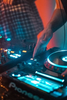 A DJ is using a modern DJ controller with colorful LED lights illuminating the scene. The hand is adjusting controls on the equipment, indicating an ongoing music mixing session. A person is partly visible in the background wearing a checkered shirt.