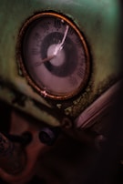 A close-up view of an old, weathered gauge with a glass cover, featuring a worn-out metallic circular frame and an aged dial showing numbers from 0 to 90. The gauge is set against a greenish, rusting background with peeling paint. A small black knob is visible beneath the gauge, suggesting a mechanical setting.