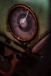 A close-up view of an old, weathered gauge with a glass cover, featuring a worn-out metallic circular frame and an aged dial showing numbers from 0 to 90. The gauge is set against a greenish, rusting background with peeling paint. A small black knob is visible beneath the gauge, suggesting a mechanical setting.