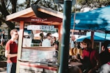 A small outdoor smoothie stand with a wooden roof and colorful signs indicating organic options. Several people gather around, some are waiting, while others are conversing or working at the stall. There's a variety of fresh produce visible, including bunches of bananas. The atmosphere suggests a market or fair, with trees and other booths in the background.