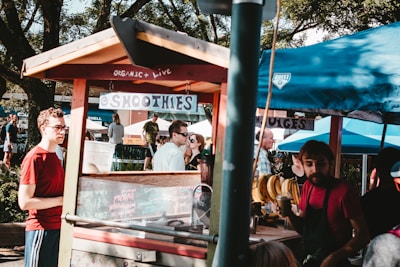 A small outdoor smoothie stand with a wooden roof and colorful signs indicating organic options. Several people gather around, some are waiting, while others are conversing or working at the stall. There's a variety of fresh produce visible, including bunches of bananas. The atmosphere suggests a market or fair, with trees and other booths in the background.