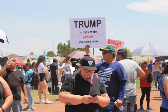 A group of people is gathered outdoors, participating in a protest. One person holds a sign with a message directed at Trump. Others are interacting, taking photos, or holding umbrellas. The setting appears to be a grassy, open area under a clear sky.