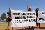 Three individuals hold a large white banner with bold black text that reads 'WHITE SUPREMACY BORDER PATROL I.C.E, CBP & EPPO' in an outdoor setting. The sky is clear and blue, and there are trees and grass visible in the background. The individuals appear to be participating in a protest or demonstration.