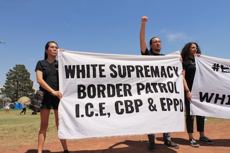 Three individuals hold a large white banner with bold black text that reads 'WHITE SUPREMACY BORDER PATROL I.C.E, CBP & EPPO' in an outdoor setting. The sky is clear and blue, and there are trees and grass visible in the background. The individuals appear to be participating in a protest or demonstration.