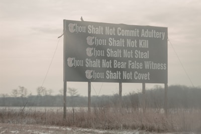 A large outdoor sign displays several commandments such as not committing adultery, not killing, not stealing, not bearing false witness, and not coveting. The background consists of an overcast sky and barren winter landscape with some sparse trees and grass.