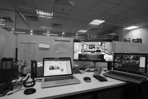 A modern office workspace with three computer screens. The center screen displays an interior design program, while the left screen shows a social media site. The right screen appears to have multiple camera feeds open. Several office items, including a telephone, notepads, coffee mug, and cables, are scattered across the desk. The office has a drop ceiling with fluorescent lighting and a transparent partition wall in the background.