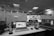 A modern office workspace with three computer screens. The center screen displays an interior design program, while the left screen shows a social media site. The right screen appears to have multiple camera feeds open. Several office items, including a telephone, notepads, coffee mug, and cables, are scattered across the desk. The office has a drop ceiling with fluorescent lighting and a transparent partition wall in the background.
