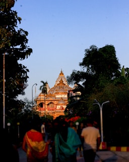 A beautifully ornate temple with intricate designs is illuminated by the golden glow of the sunset. Surrounded by lush greenery and trees, the temple features a detailed domed structure. People walk along a path towards the temple, some dressed in traditional attire, creating a serene atmosphere.
